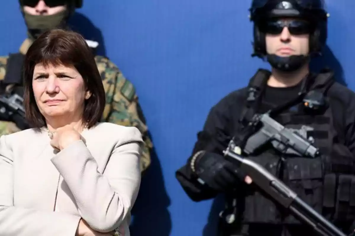 Una mujer de cabello oscuro y chaqueta clara está de pie junto a dos personas vestidas con uniformes tácticos y armadas, sobre un fondo azul.