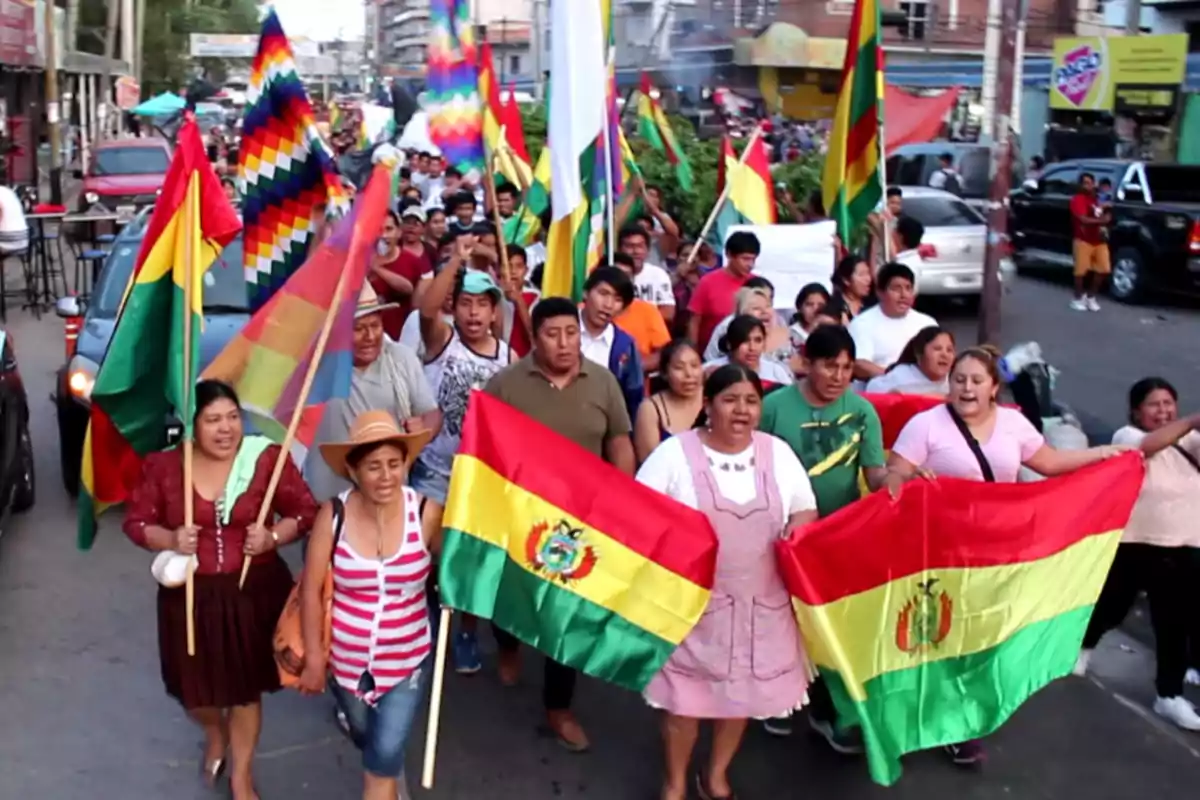 Un grupo de personas marcha por una calle llevando banderas de Bolivia y la bandera Wiphala.