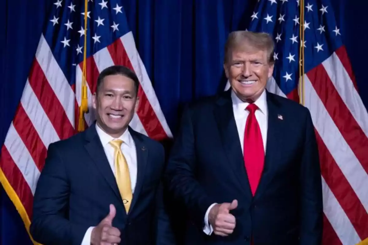 Dos personas sonriendo y levantando el pulgar frente a banderas de Estados Unidos.