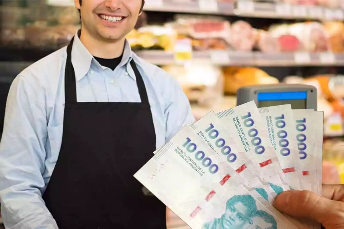 Un hombre con delantal negro sonríe detrás de un mostrador mientras una mano sostiene billetes de 10,000 pesos argentinos.