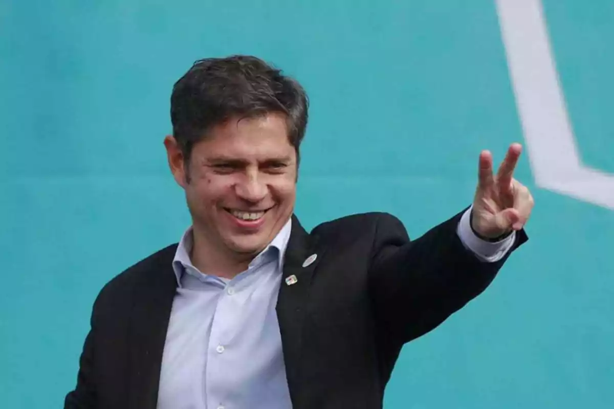 Un hombre sonriente con traje oscuro y camisa clara hace un gesto de paz con la mano frente a un fondo azul.