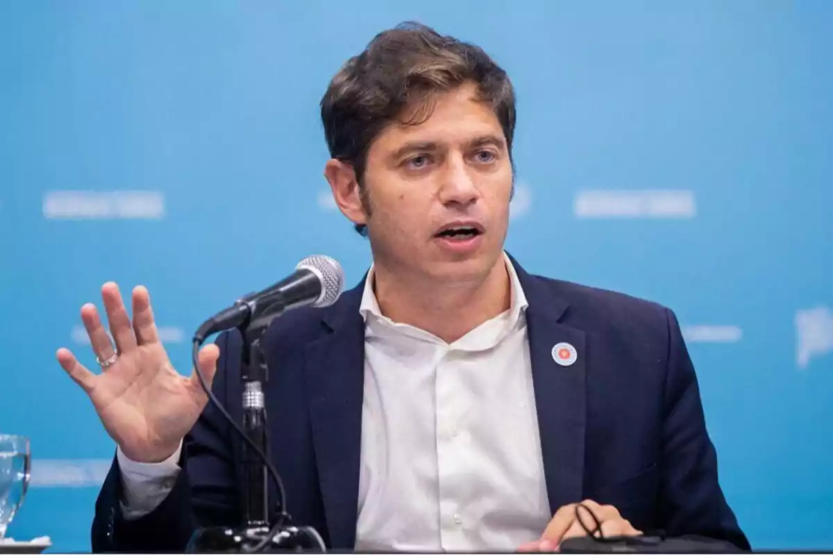 Hombre hablando en una conferencia de prensa con un micrófono frente a él y fondo azul.