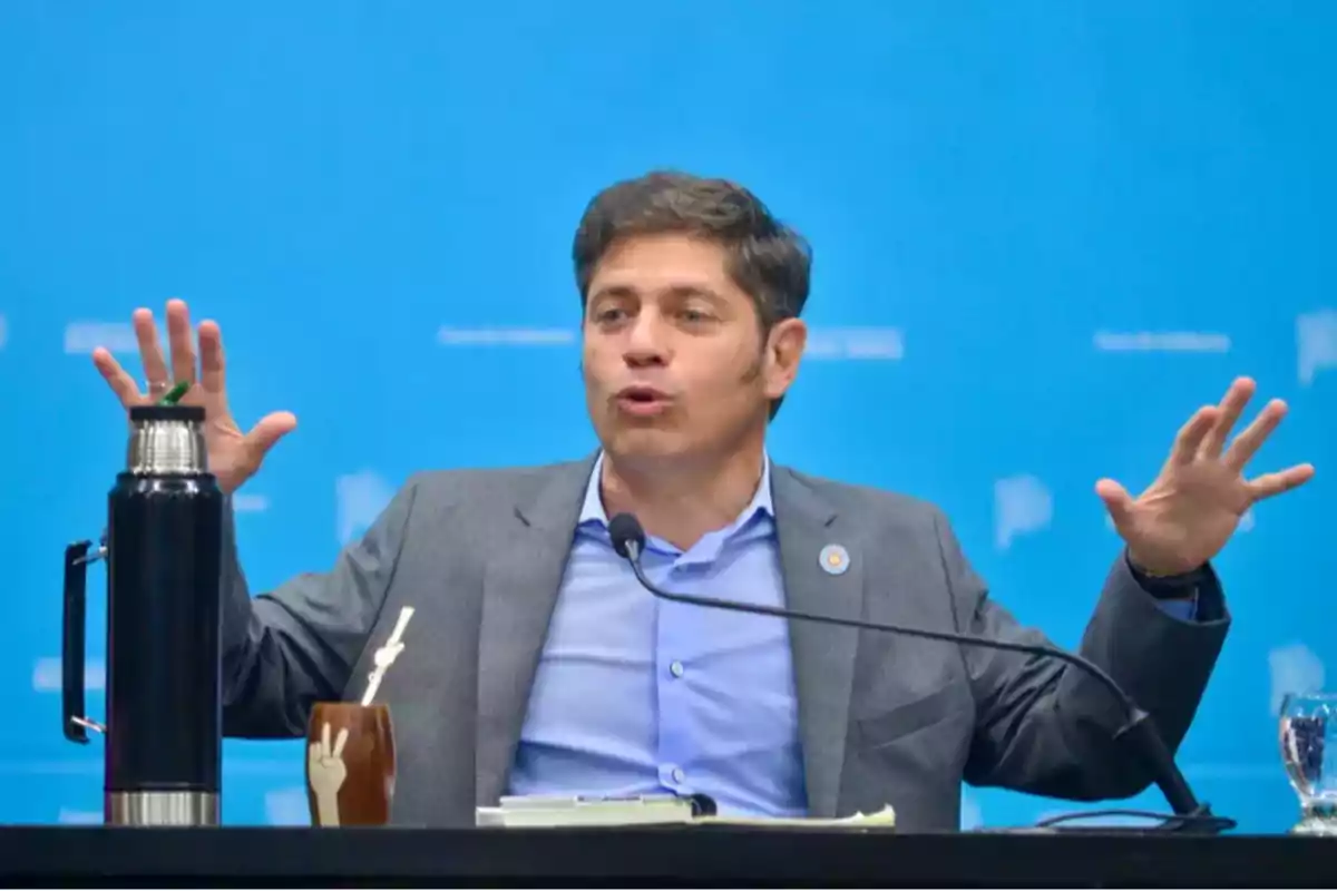 Un hombre en traje gesticula con las manos mientras habla en una conferencia con un fondo azul y un termo y mate sobre la mesa.