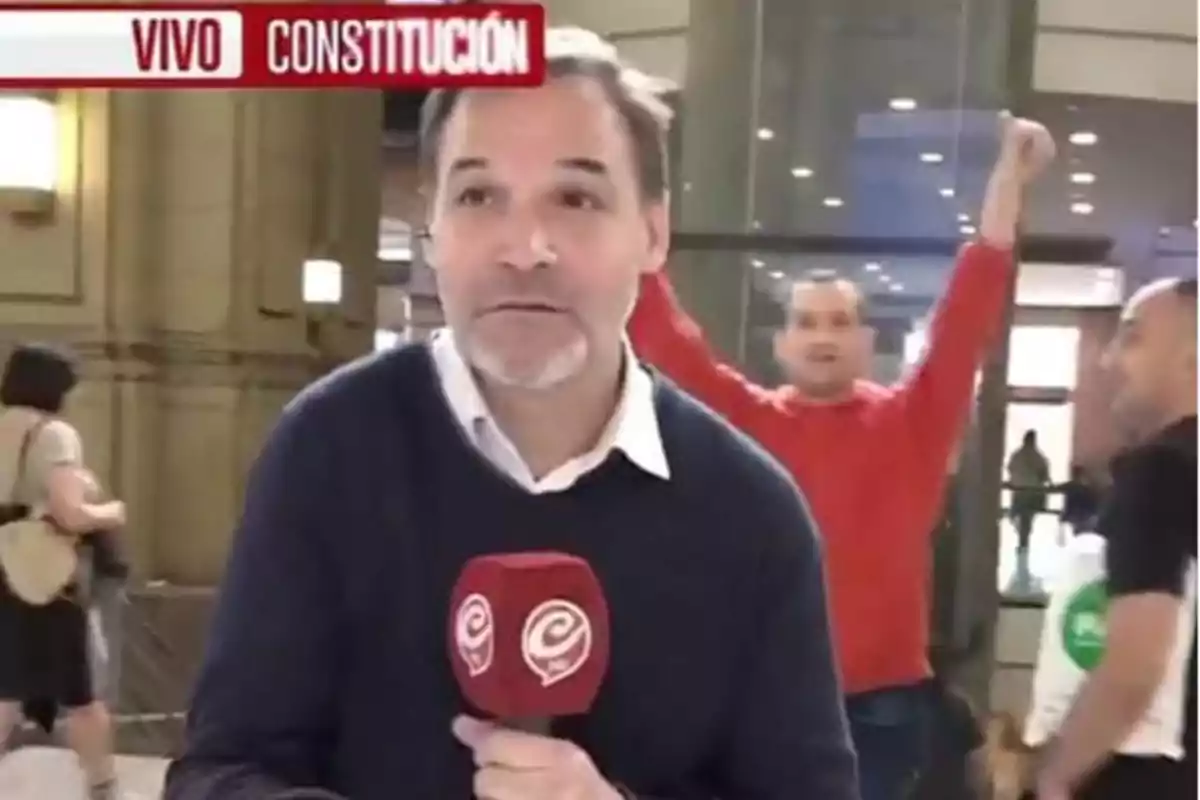 Un reportero de televisión está transmitiendo en vivo desde la estación de Constitución mientras una persona en el fondo levanta los brazos.