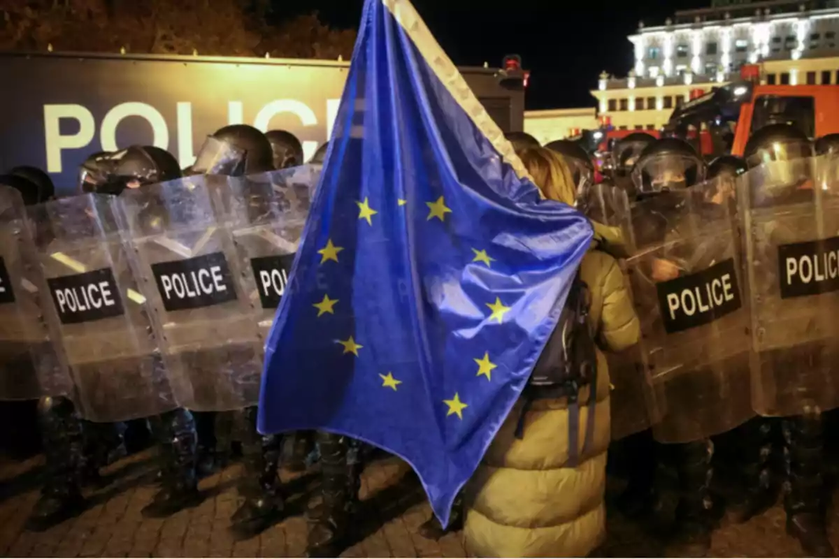 Una persona sostiene una bandera de la Unión Europea frente a una fila de policías con escudos en una manifestación nocturna.