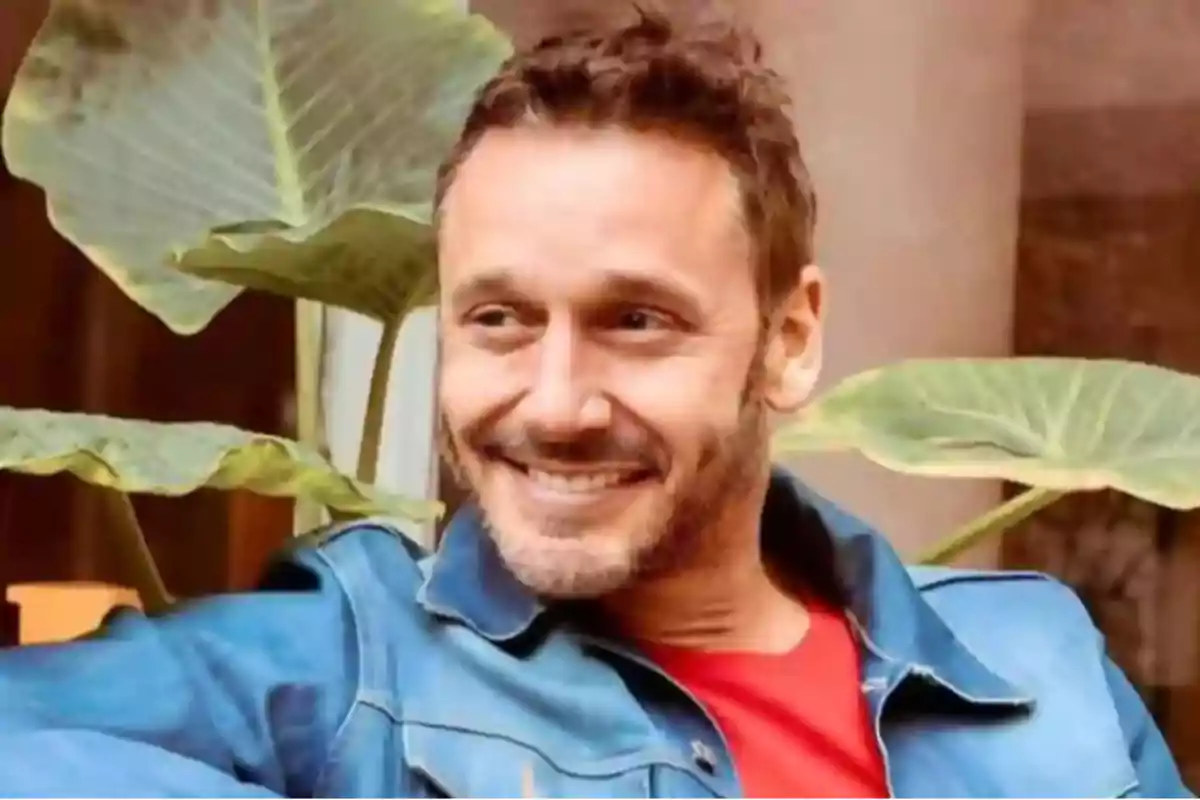 Un hombre sonriente con barba y cabello corto lleva una chaqueta de mezclilla y una camiseta roja con hojas grandes de planta en el fondo.