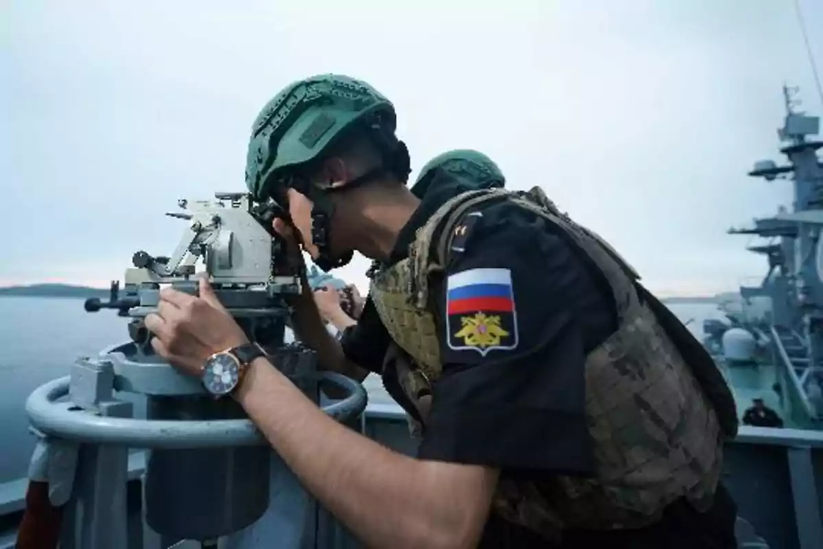 Un militar con casco y chaleco antibalas observa a través de un dispositivo óptico en la cubierta de un barco, con el mar y otro barco al fondo.