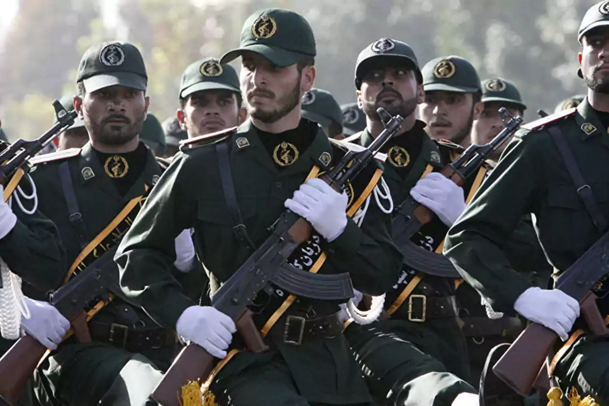 Un grupo de soldados uniformados marchando en formación, portando rifles y vistiendo gorras verdes con insignias.