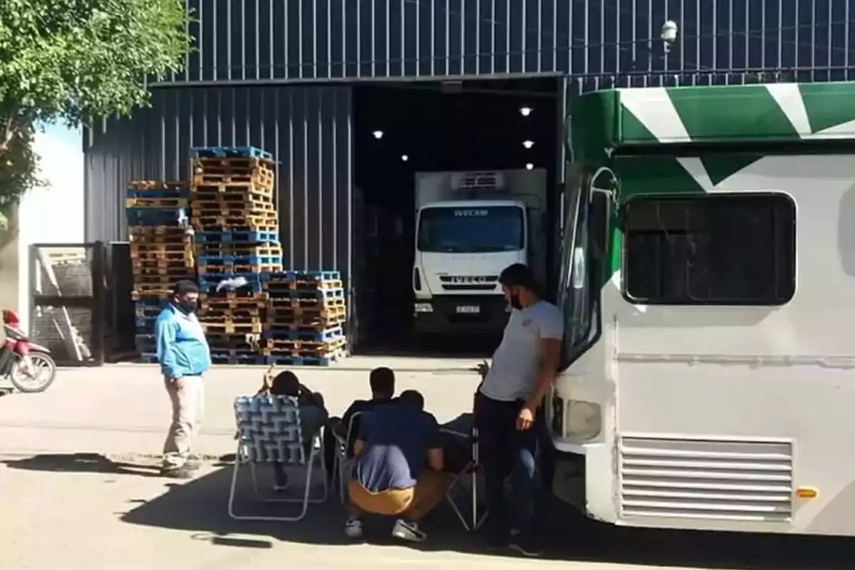 Varias personas descansan afuera de un almacén con palets apilados y un camión estacionado en la entrada.