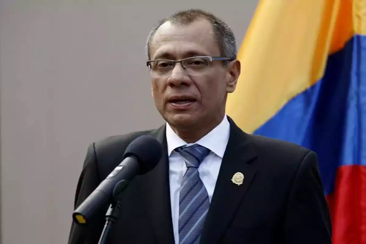 A man with glasses and a dark suit speaks in front of a microphone with a flag in the background.