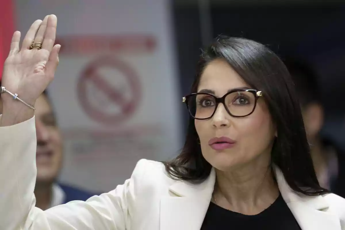 Mujer con gafas levantando la mano en un entorno interior.