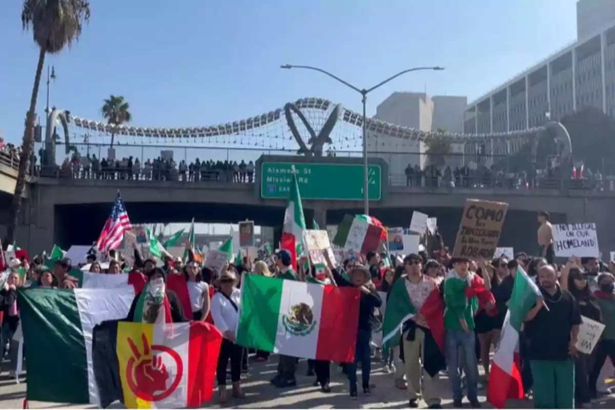 Un grupo de personas participa en una manifestación, llevando banderas de México y Estados Unidos, así como pancartas, mientras caminan por una calle con un puente peatonal y un letrero de tráfico en el fondo.