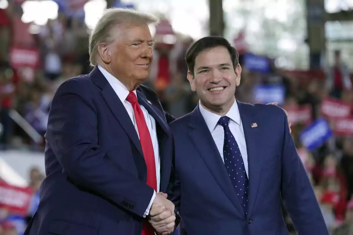 Two men in dark suits shake hands and smile at an event with an audience in the background.