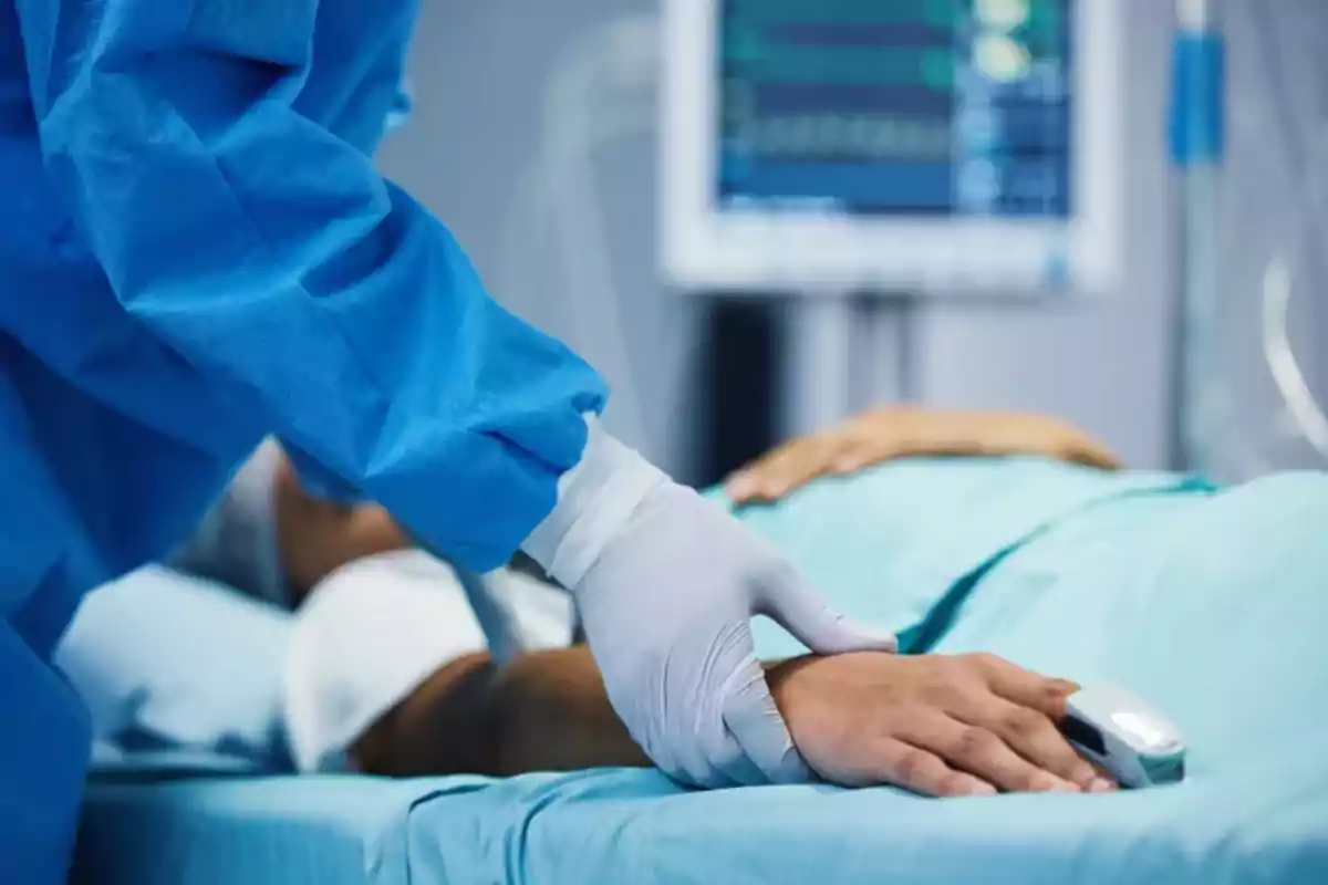 Un profesional de la salud con guantes sostiene la mano de un paciente en una cama de hospital, con un monitor médico en el fondo.