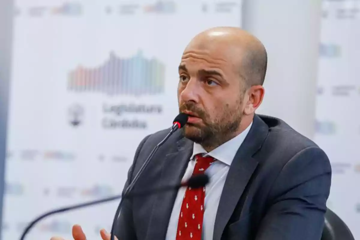 Un hombre calvo con barba hablando en un micrófono durante una conferencia de prensa, con un fondo que muestra un cartel de la Legislatura de Córdoba.