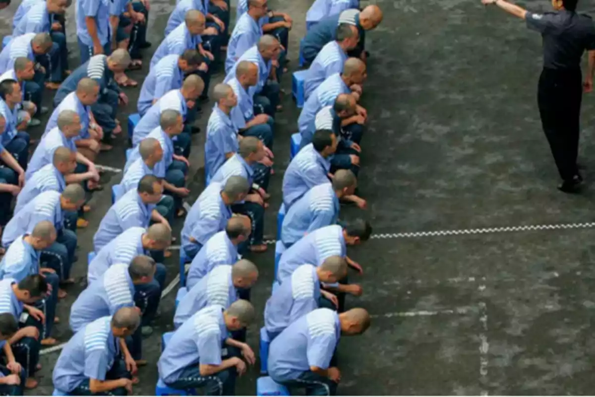 Personas sentadas en filas con uniformes azules, con la cabeza agachada, mientras una persona de pie al frente parece dar instrucciones.