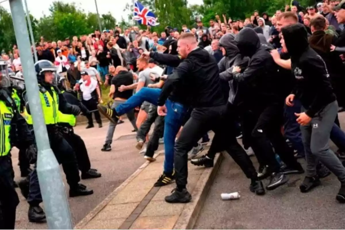 Un grupo de manifestantes se enfrenta a la policía en una protesta, con una multitud observando en el fondo y una bandera del Reino Unido ondeando.