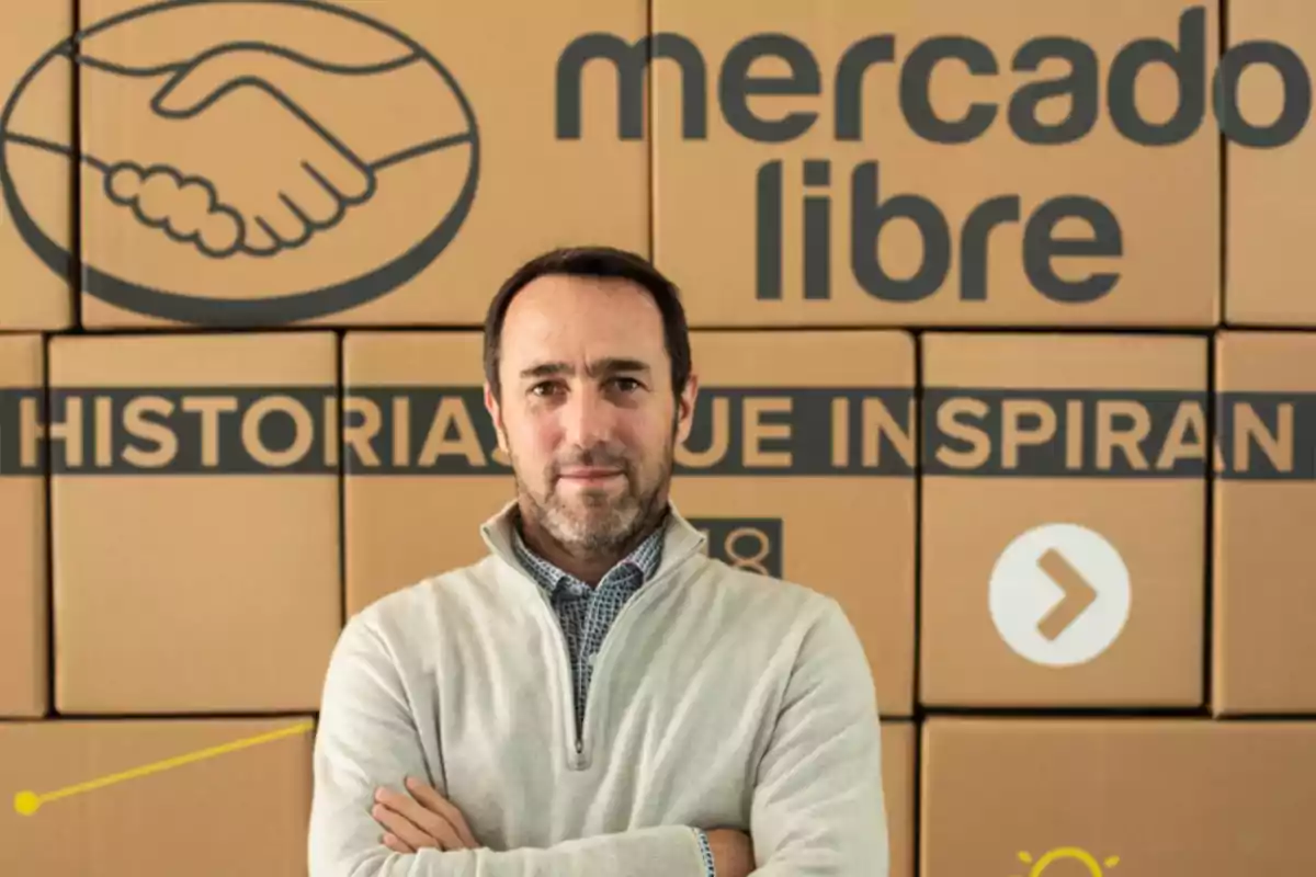 Un hombre con suéter claro y camisa de cuadros posa con los brazos cruzados frente a cajas de cartón con el logo y nombre de Mercado Libre.