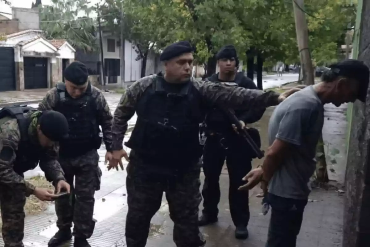 La imagen muestra a varios policías armados y uniformados deteniendo a un hombre contra una pared en una calle residencial.