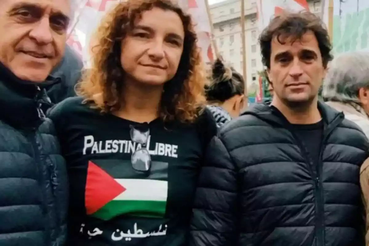 Tres personas posan juntas en una manifestación, una de ellas lleva una camiseta con la bandera de Palestina y el texto "Palestina Libre".