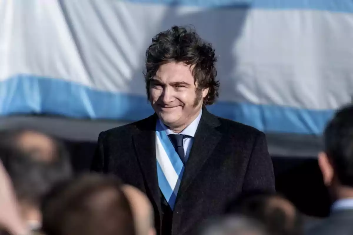 Hombre con traje oscuro y corbata azul claro frente a una bandera argentina.