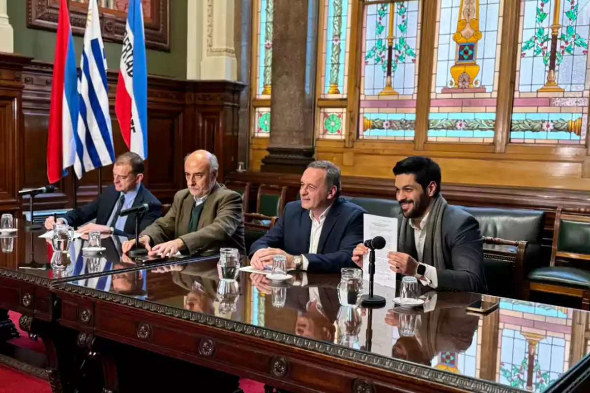 Cuatro hombres sentados en una mesa de madera oscura con micrófonos y documentos, en una sala con vitrales y banderas en el fondo.