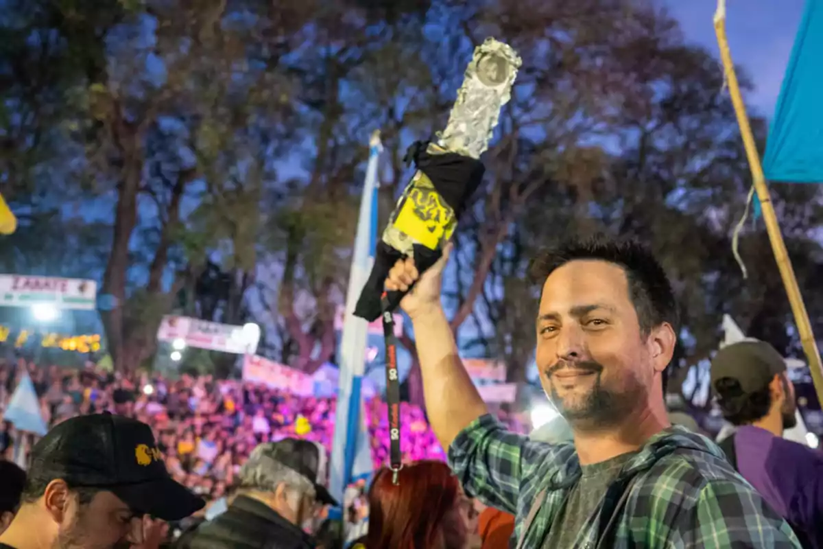 Un hombre sonriente sostiene un objeto decorado en un evento al aire libre con una multitud de personas y árboles en el fondo.