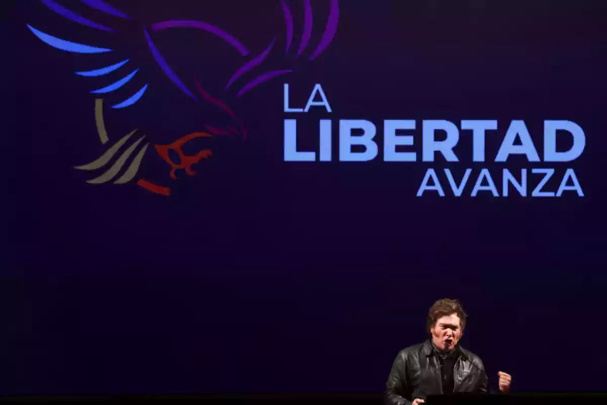 Una persona hablando en un escenario con un fondo que muestra un logo de un águila y las palabras "La Libertad Avanza".