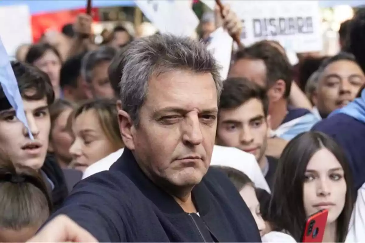 Un hombre de cabello canoso y expresión seria se encuentra rodeado de una multitud de personas, algunas de las cuales sostienen pancartas y banderas.