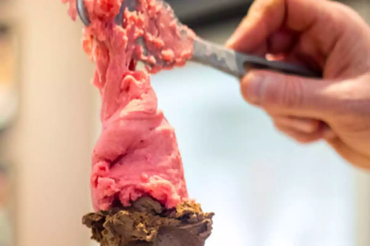 Una mano sostiene una cuchara sirviendo helado de fresa sobre una base de helado de chocolate.