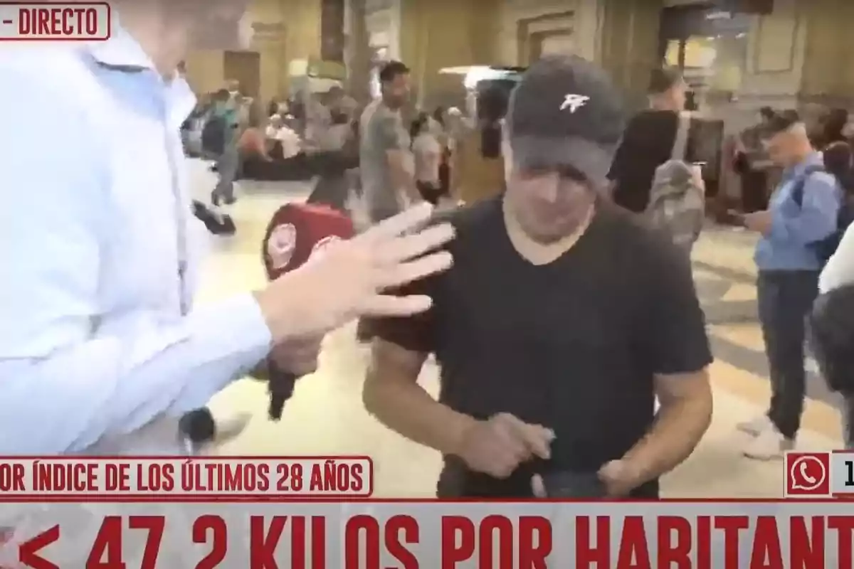 Un reportero está entrevistando a una persona en un lugar concurrido mientras sostiene un micrófono rojo con un logotipo.