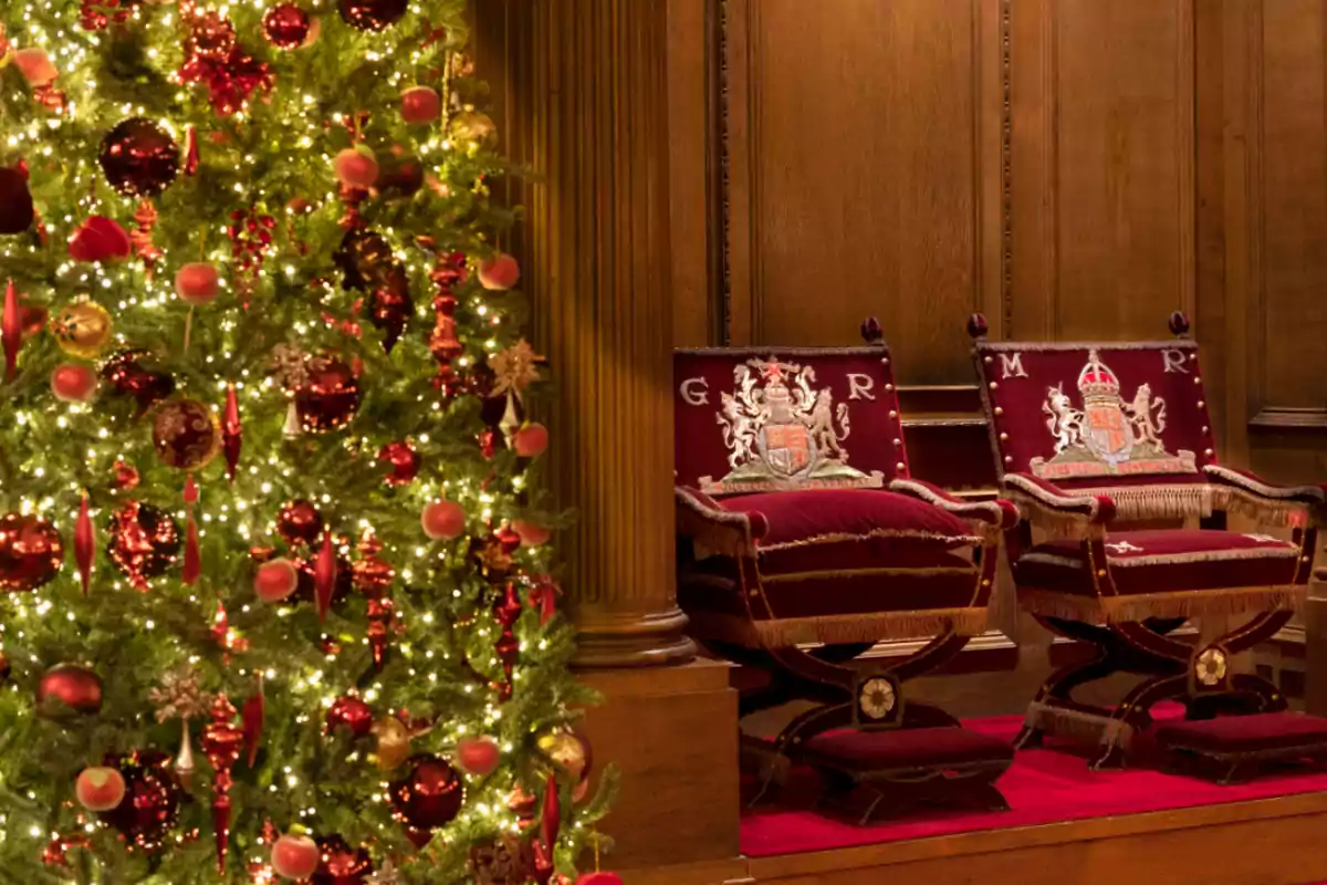 Un árbol de Navidad decorado con luces y adornos rojos junto a dos sillas elegantes con cojines bordados en una sala de madera.