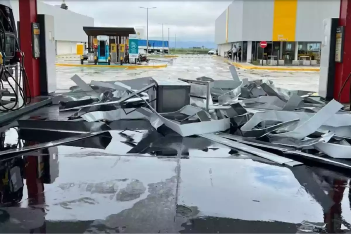 Techo colapsado en una gasolinera con escombros metálicos esparcidos en el suelo mojado.