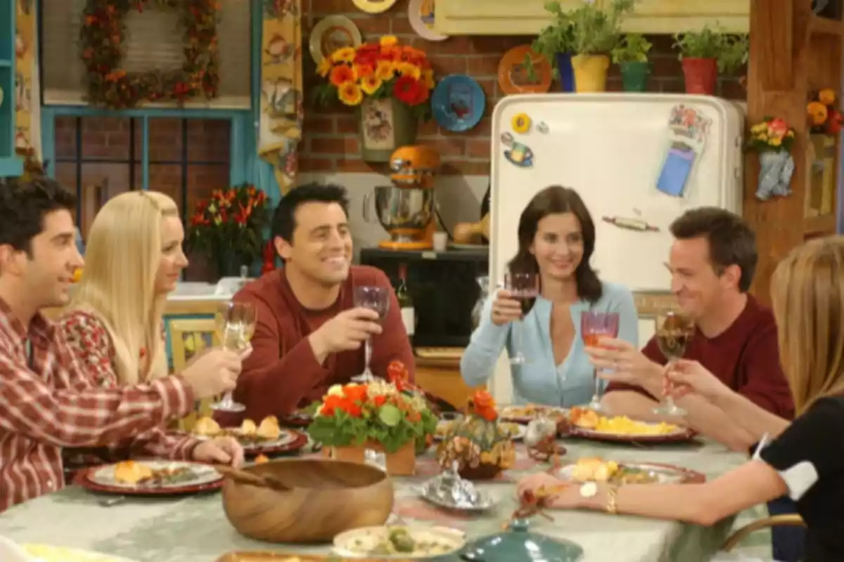 Un grupo de amigos sonríe mientras brinda alrededor de una mesa decorada con flores y comida en un ambiente hogareño.