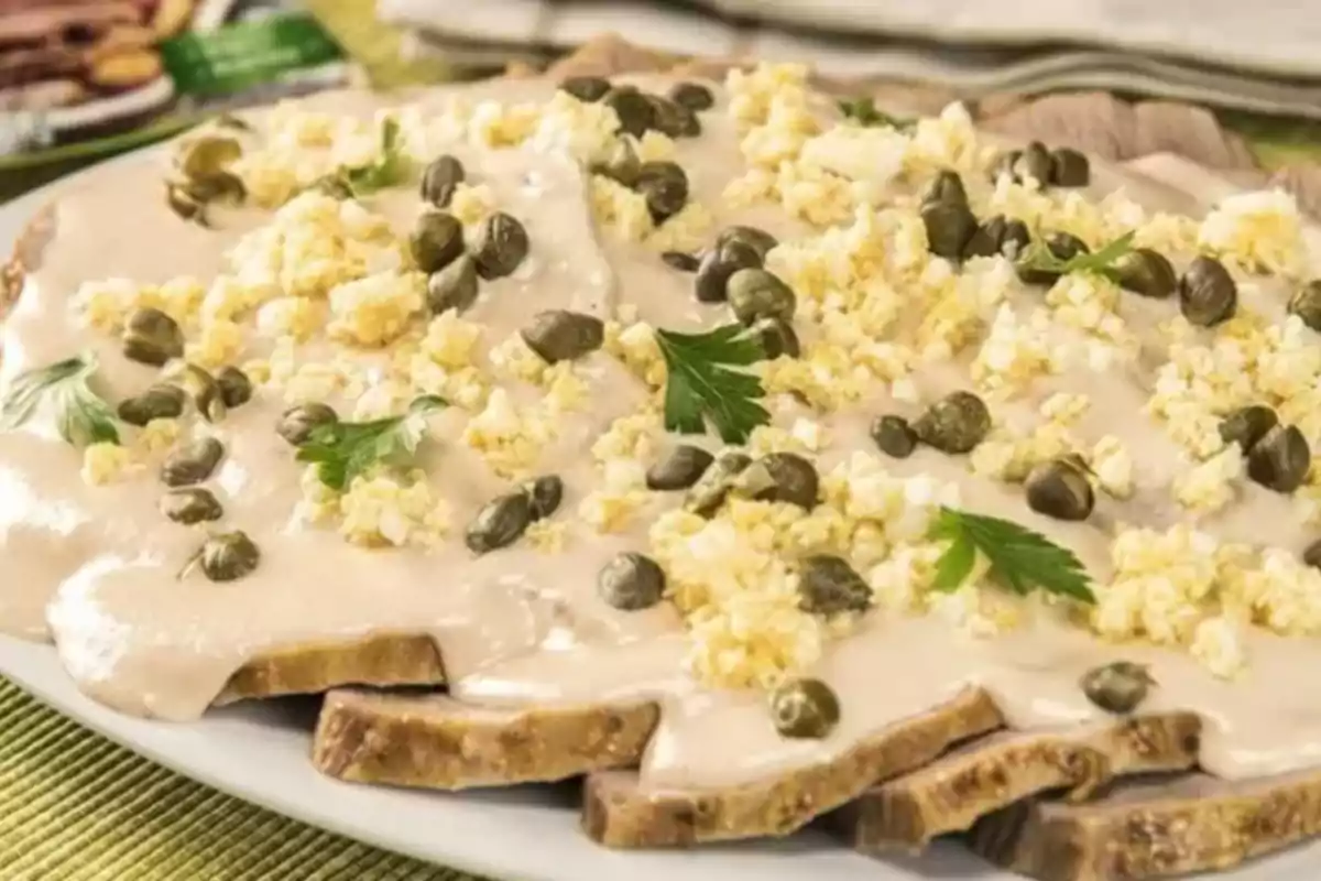 Plato de vitel toné con salsa cremosa, alcaparras y perejil sobre un plato blanco.