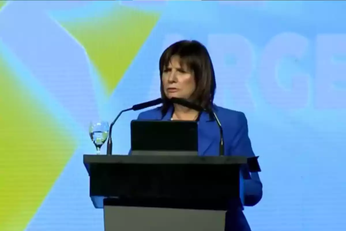 Una persona en un podio hablando en un evento con un fondo azul y amarillo.