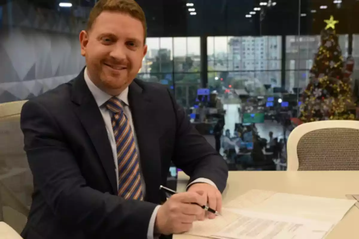 Hombre sonriendo mientras firma documentos en una oficina moderna con un árbol de Navidad al fondo.