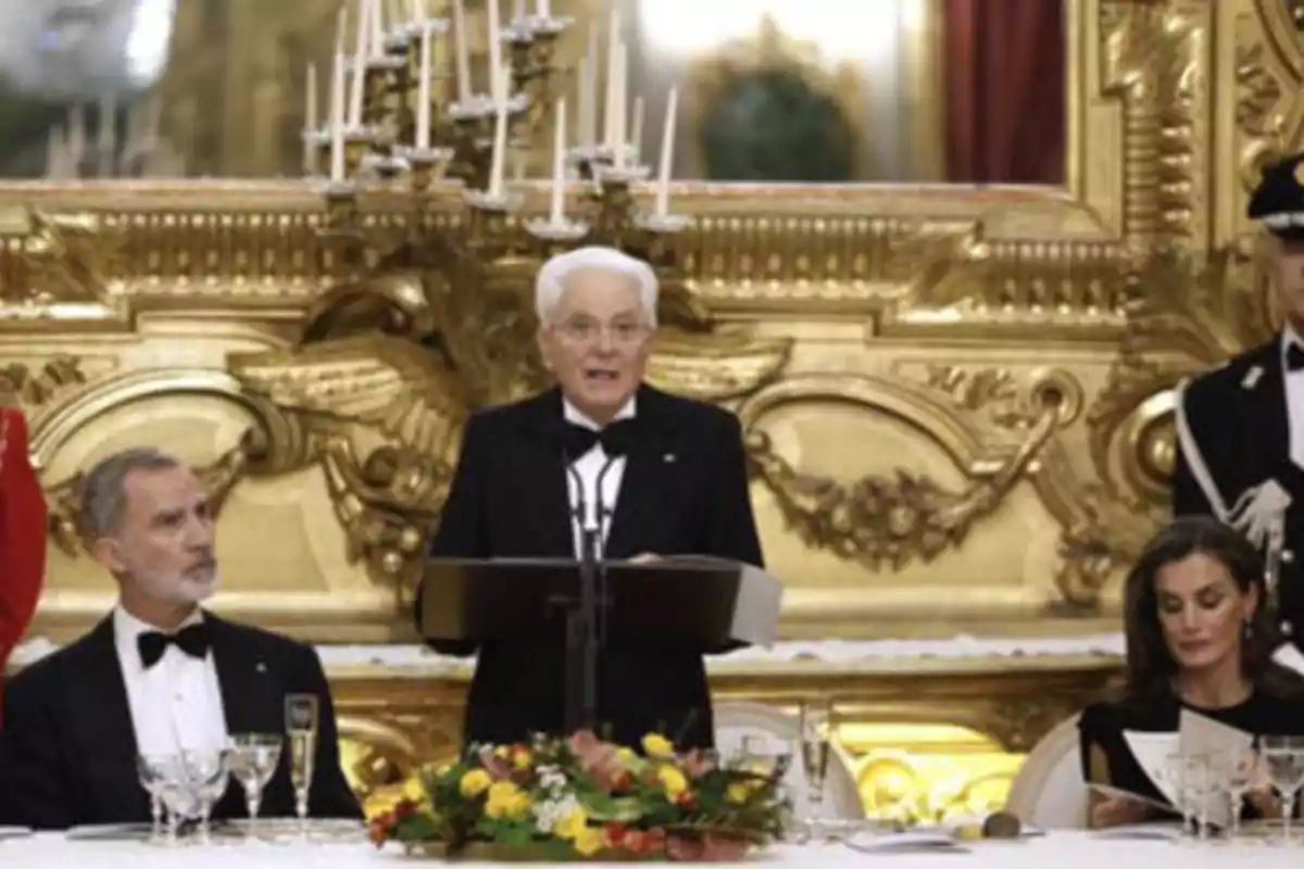 Un hombre mayor de pie hablando en un podio en un evento formal con personas sentadas a su alrededor en una sala decorada con detalles dorados y candelabros.