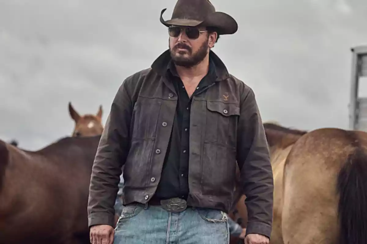 Hombre con sombrero de vaquero y gafas de sol, vestido con chaqueta marrón y jeans, rodeado de caballos.