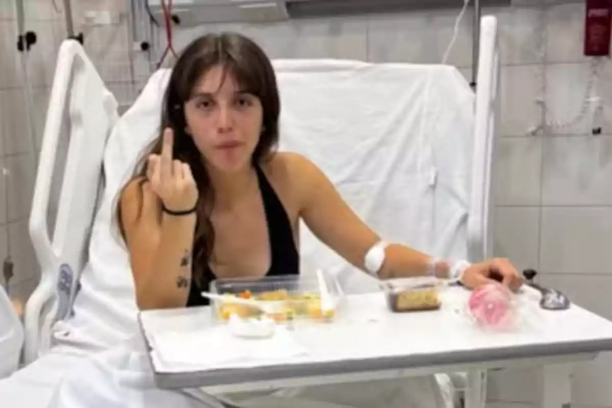 Una persona en una cama de hospital haciendo un gesto con la mano mientras tiene una bandeja de comida frente a ella.