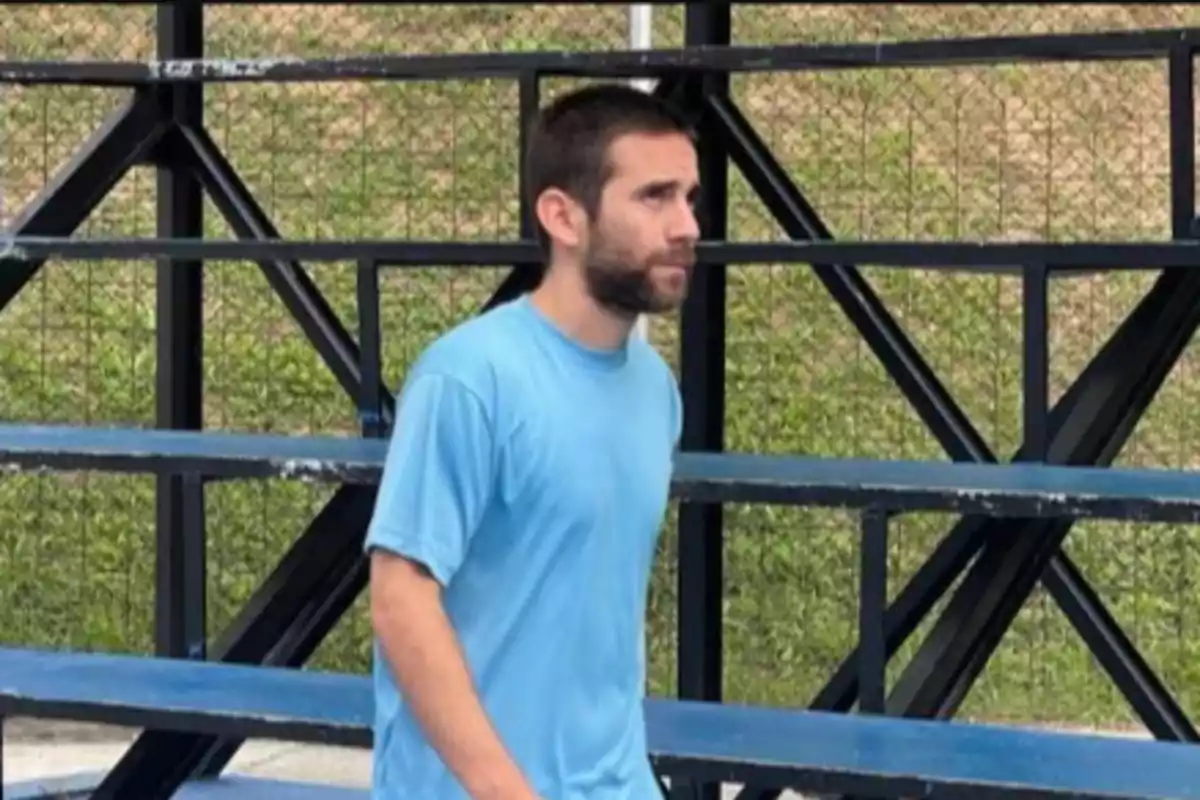 Hombre con camiseta azul caminando frente a unas gradas metálicas.