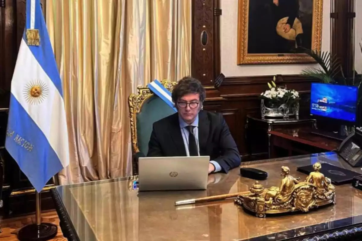 Un hombre con traje y gafas trabaja en una computadora portátil en un escritorio elegante con una bandera argentina a su lado.