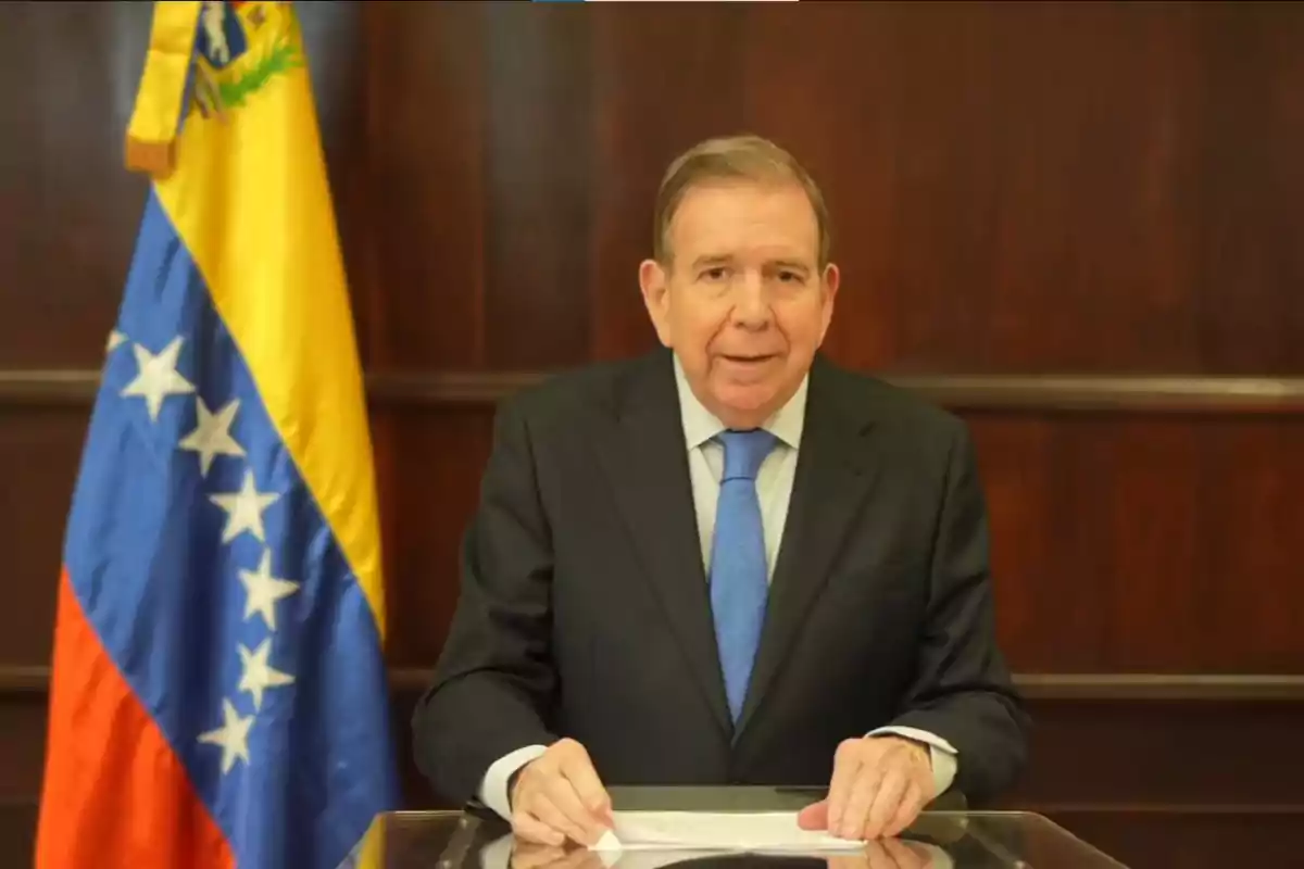 Un hombre de traje oscuro y corbata azul está de pie frente a un podio con una bandera de Venezuela a su lado.