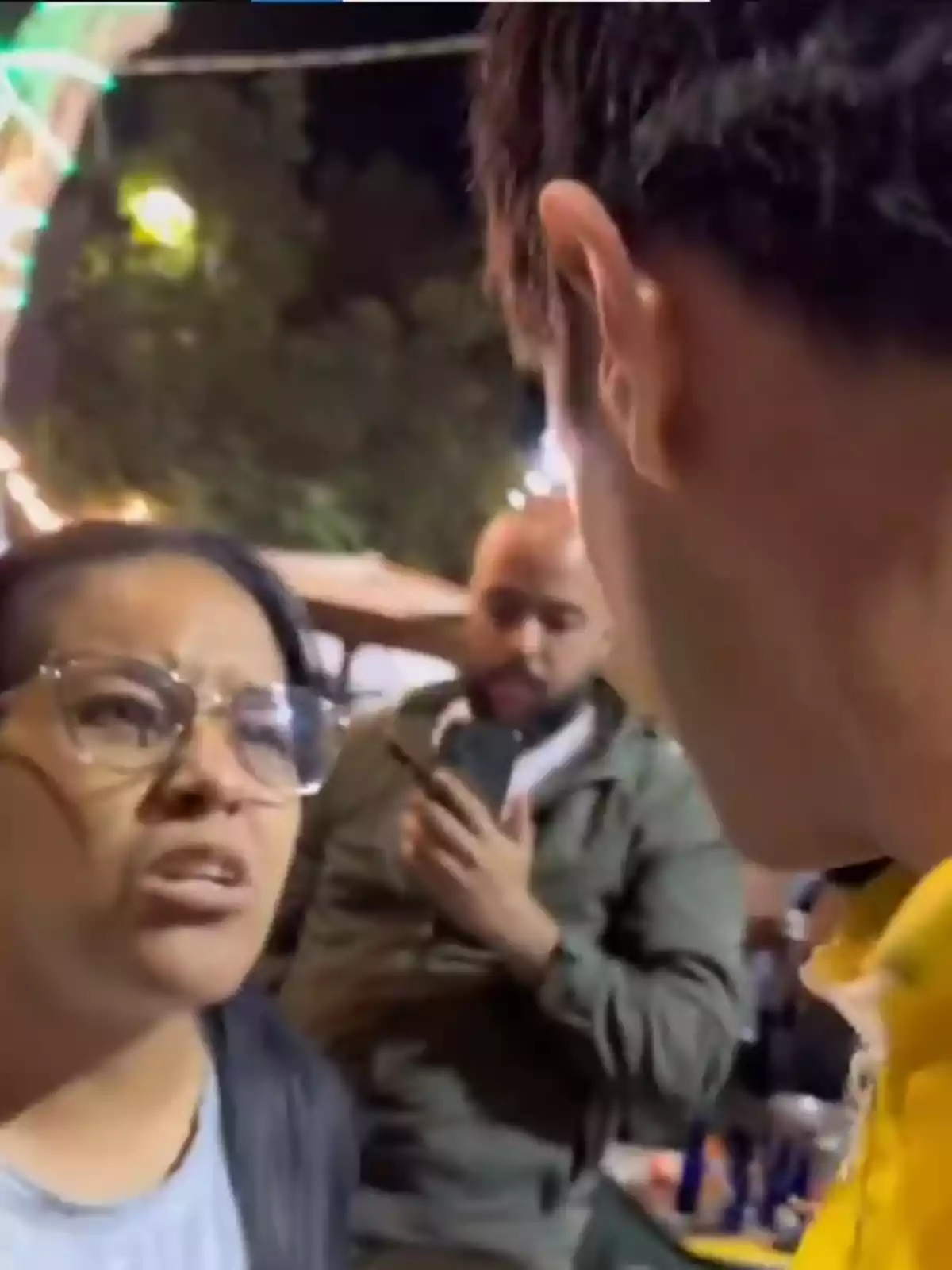 Una mujer con gafas parece estar discutiendo con un hombre mientras otra persona graba la escena con su teléfono en un entorno nocturno al aire libre.