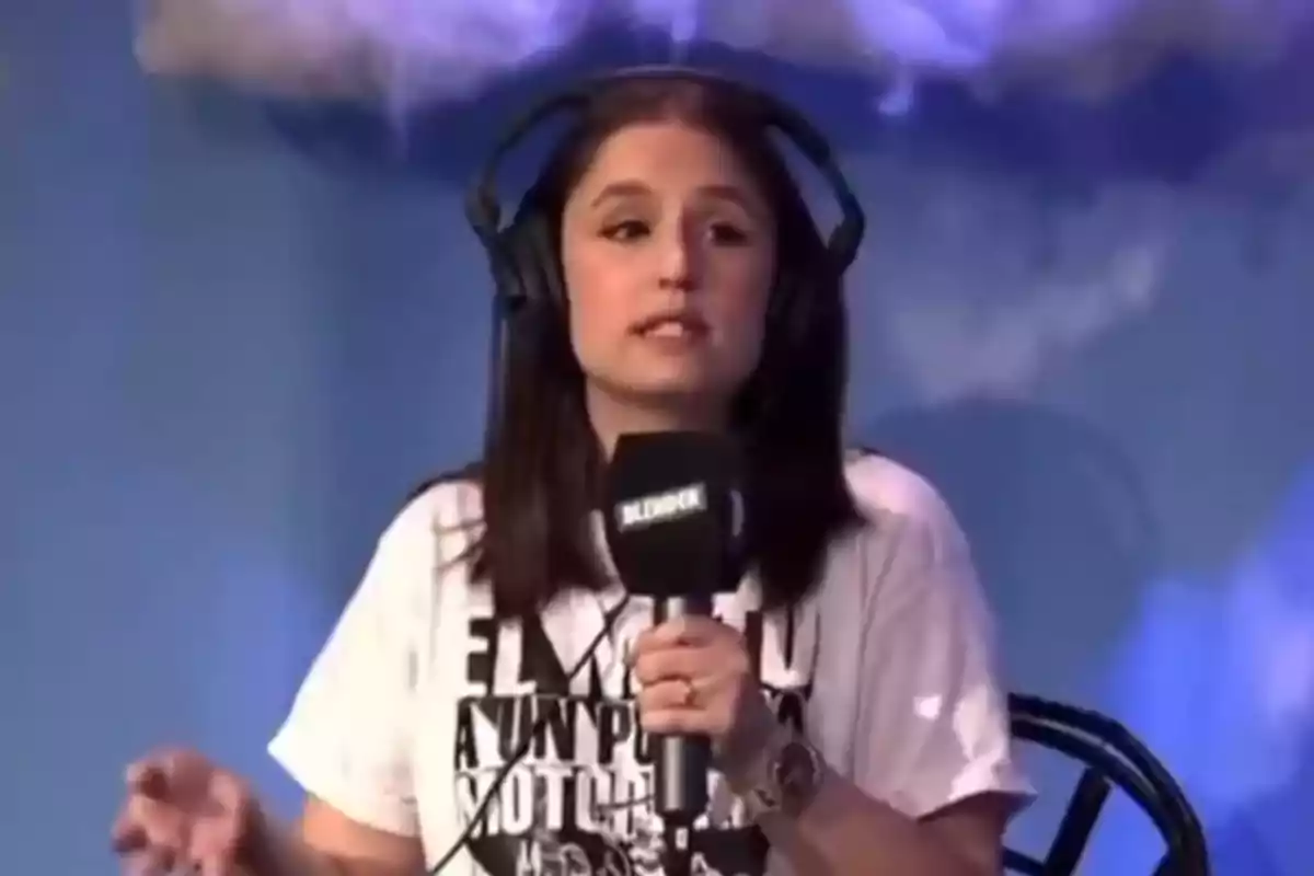 Una persona con auriculares y camiseta blanca habla frente a un micrófono en un estudio con fondo azul.