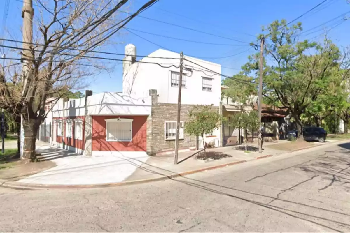 Edificio de dos pisos en una esquina con fachada de ladrillo rojo y blanco rodeado de árboles y cables eléctricos.