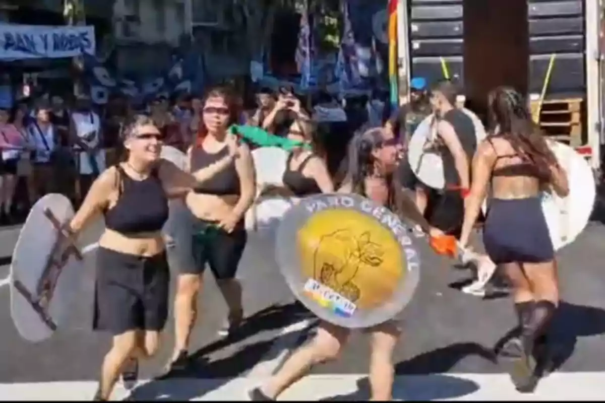 Un grupo de personas participa en una manifestación en la calle, algunas llevan escudos y pancartas, mientras otras observan desde el fondo.