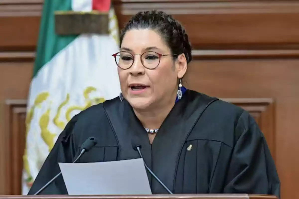 Una persona con gafas y vestimenta formal habla frente a un micrófono con una bandera de México de fondo.