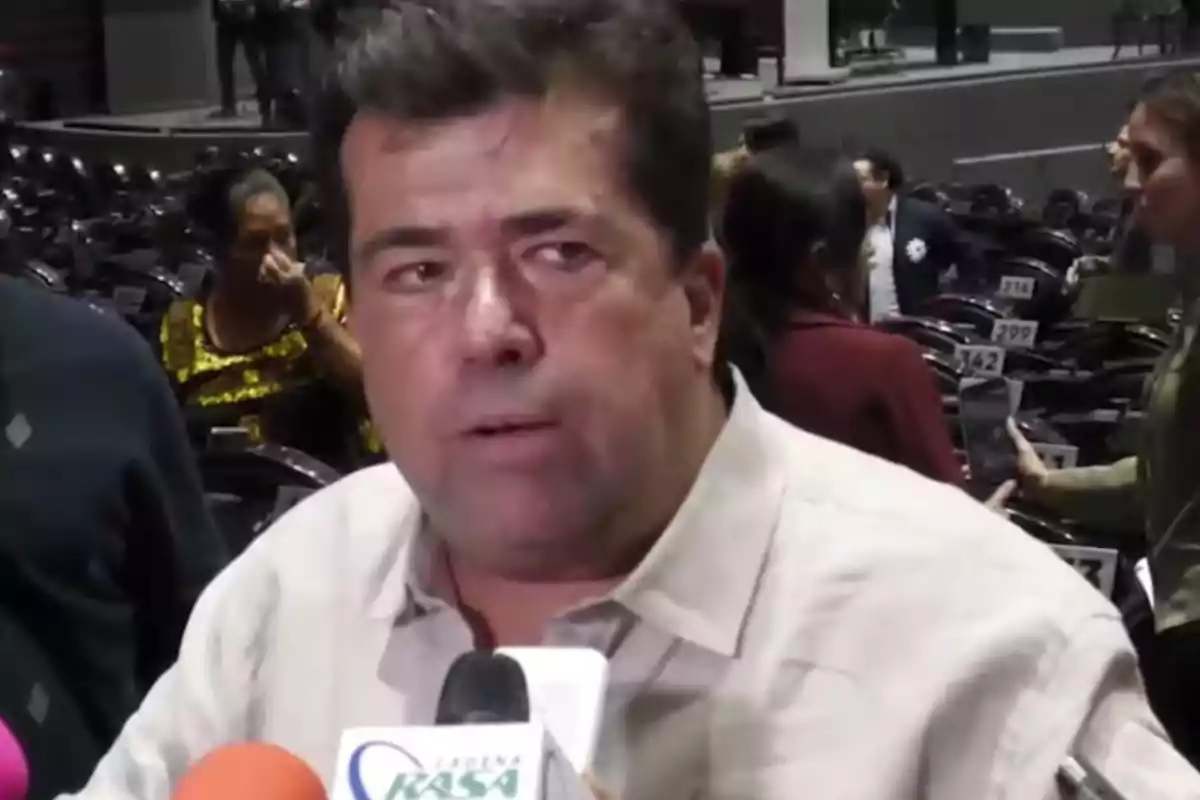 A man speaking in front of several microphones at an event with people in the background.