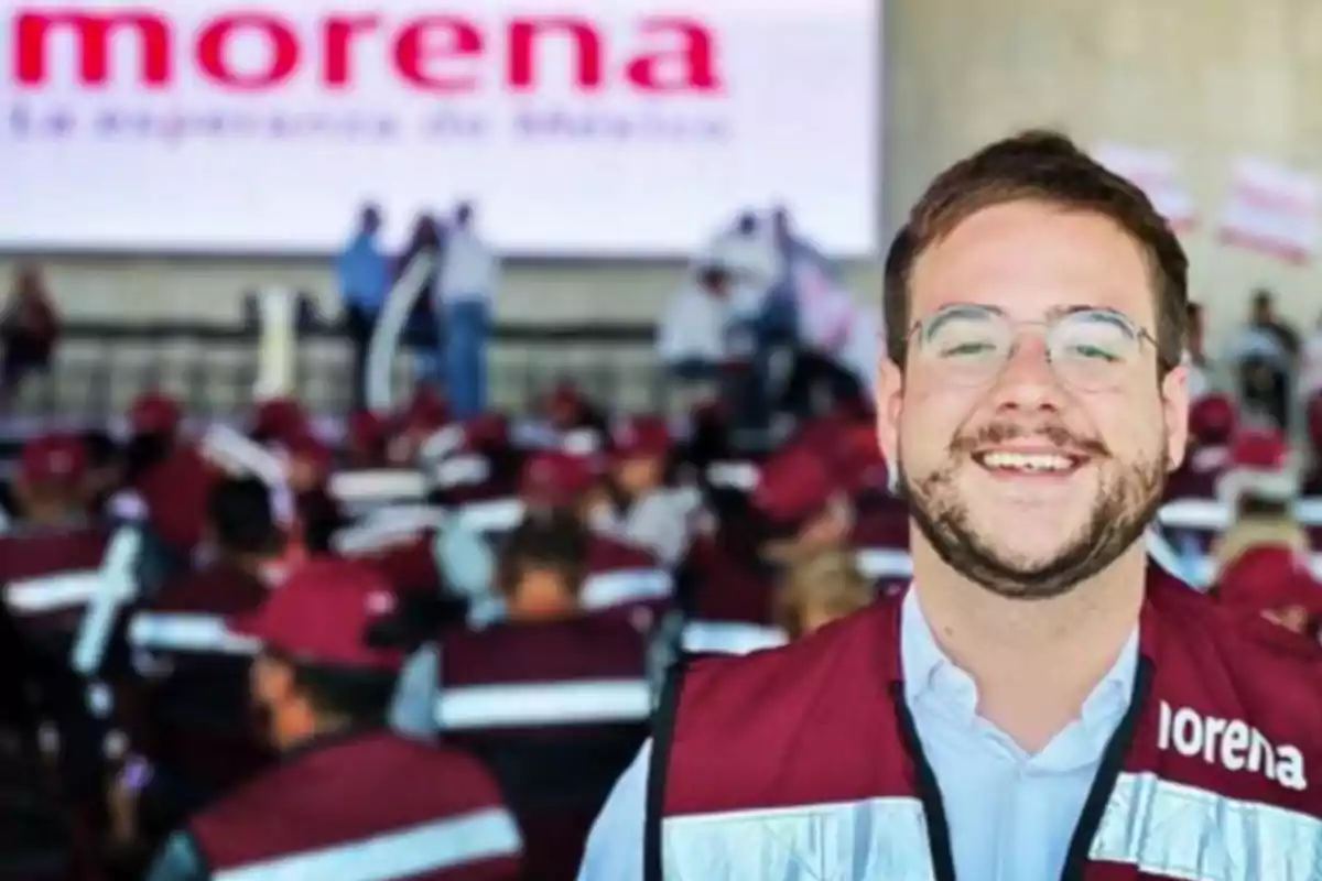 A smiling man in a red vest and glasses at a Morena event with a group of people in the background.